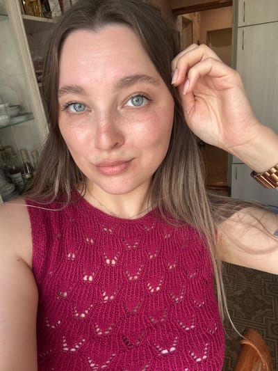 The image is a close-up, waist-up portrait of a young woman with long, blonde hair and blue eyes. She is looking directly at the camera, with her right hand touching her forehead and hair. She is wearing a bright pink, sleeveless, knitted top with a V-neck and an intricate lace-like pattern. A delicate silver necklace with a small pendant is visible around her neck. On her right wrist, she is wearing a rose gold watch. The background is blurred but appears to be an indoor setting, with shelves containing bottles and jars on the left, and a plain wall with cabinetry on the right. The lighting suggests it is daytime. There is no indication of the specific location within Yaroslavka, Russia.