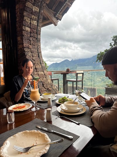 Captured indoors at a restaurant in Sa Pa Ward, Vietnam, the image presents a scene from a dining table. A woman with braided hair, wearing a black shirt, is engrossed in her phone while holding a straw in a glass of orange juice. Next to her is a plate with what appears to be a tomato salad. At the same table, another individual partially visible at the top, dressed in a light brown sweater, is also occupied with a phone. The table holds assorted dishes, including salad bowls and empty plates, along with silverware. The background reveals an expansive view of cloudy skies and a mountain range through a glass window, complemented by the restaurant's brick arch structure.