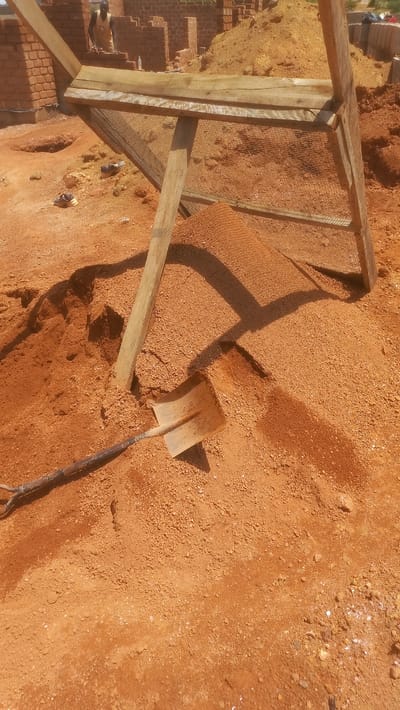 A construction site in Katon Rikkos, Nigeria, is depicted in this image. The scene is dominated by a large pile of reddish-orange soil, likely sand or gravel, which has been sifted through a wooden sieve supported by two vertical wooden posts. A shovel with a worn metal blade and a wooden handle rests partially buried in the soil, its tip pointing upwards. In the background, unfinished brick structures are visible, suggesting ongoing construction work. A person wearing a yellow garment and dark shorts stands near the brickwork, appearing to be involved in the construction. The ground is uneven and covered in loose soil and small stones, with some scattered debris, including a pair of worn athletic shoes. The lighting suggests it is daytime, with shadows cast by the sieve and other objects on the ground.