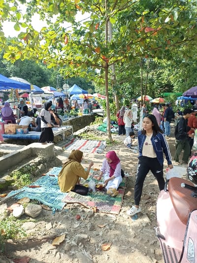 This image depicts a vibrant outdoor market or social gathering in Mataram, Indonesia, bustling with people under the bright daylight. The scene is set amidst abundant green foliage and trees, which provide natural shade over the area.
In the foreground, two women are seated on colorful woven mats on the dirt ground, appearing to enjoy a meal or picnic. Food items are visible before them. To their right, a young woman stands and walks towards the right of the frame, carrying a white plastic bag. She is wearing a navy blue jacket with white stripes (displaying an Adidas logo and text) over a light-colored top, and black pants with a Reebok logo. Partially visible on the far right is the front part of a pink scooter.
The midground and background reveal numerous market stalls sheltered by various colorful canopies and umbrellas, including shades of blue, red, and white. Many people, including several women wearing hijabs, are seen walking, standing, and congregating around these stalls, suggesting buying, selling, and socializing activities. Some individuals are seated on raised platforms next to stalls.
The ground consists of dirt and gravel paths, with some concrete structures possibly delineating walkways or drainage. The strong sunlight casts clear shadows, indicating a sunny day, likely during the late morning or early afternoon. The overall atmosphere appears relaxed and social, characteristic of a lively community market or public gathering in a tropical setting. No specific product names or detailed texts are clearly legible on the market stalls.