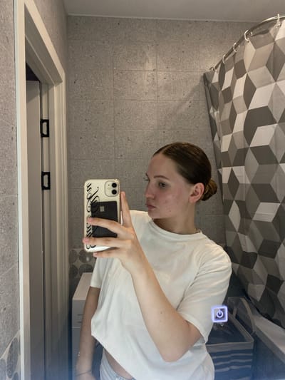 A young woman is taking a mirror selfie in a bathroom. She is wearing a white t-shirt and has her hair pulled back in a bun. Her face is turned to the side, and she is holding a smartphone with a case that has a cardholder in it. The bathroom has grey tiled walls and a shower curtain with a geometric pattern. In the background, a laundry basket can be seen.