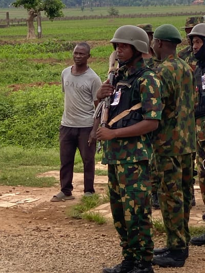 A low-angle, outdoor shot shows a single African male, wearing a grey t-shirt and dark trousers, holding an AK-47 rifle. He is positioned in front of two soldiers wearing camouflage fatigues and helmets. The area is a gravel patch next to a grassy field. A row of trees and a fenced area are visible in the background. The location is in the city of Maigizo, Nigeria.