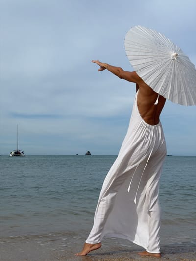 A person stands on a beach in Baan Plai Laem, Thailand, wearing a long, flowing white dress and holding a white umbrella over their shoulder. The person's back is to the viewer, with their left arm extended to the left, palm facing away. They are barefoot and appear to be walking or posing at the water's edge, with gentle waves lapping at their feet and the hem of their dress. The ocean stretches out before them, calm and blue-grey, with a distant sailboat and a small rock formation visible on the horizon. The sky is a pale blue with wispy white clouds. The overall scene is serene and evokes a sense of peacefulness.