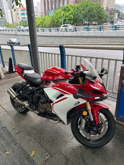 The image depicts a red and white motorcycle parked on a tiled sidewalk. The motorcycle is next to a metal railing and a grey support column. In the background, there is a street with cars and trees, and buildings are visible in the distance. The location is 龙溪街道, China.
