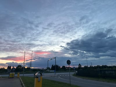 The image captures a late evening sky with dramatic cloud formations over the town of Laagri alevik, Estonia. The sky is filled with a mix of gray and dark storm clouds, punctuated by streaks of vibrant pink and orange from the setting sun. In the foreground, a paved area transitions into a grassy verge, bordering a road. Visible on the paved area are two yellow structures, likely payment or information kiosks, and a black trash receptacle. Several lampposts and traffic signs are present along the road, which appears to be a junction or intersection. A line of trees and some buildings are visible in the mid-ground, silhouetted against the colorful sky. The overall impression is a serene yet dramatic natural scene with hints of urban infrastructure.