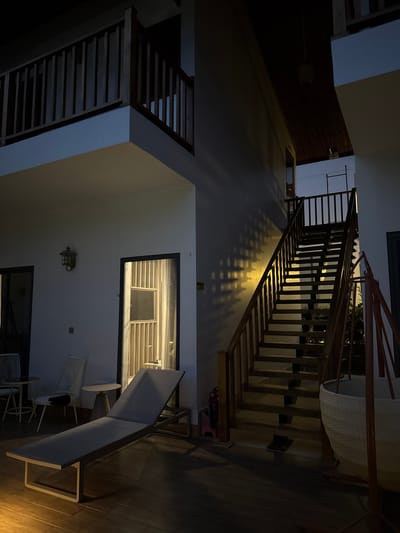 This media file captures a multi-level interior space, likely within a resort, hotel, or large residence in Phú Quốc, Vietnam, during the evening or night. The scene is dominated by a large wooden staircase ascending from the mid-ground towards the upper right. A light source positioned beneath this staircase casts dramatic, patterned shadows of the steps and balusters onto the adjacent white wall, creating a striking visual effect. The staircase features dark wooden treads and railings, with open risers.

On the ground floor, a long, modern lounge chair with light-colored upholstery is positioned on a dark wooden floor, next to two small, round white tables and matching chairs. Further into the ground floor area, another white-painted staircase with dark wooden treads and railings is visible, leading to a second-floor landing on the left. A rectangular opening or doorway on the ground floor wall reveals a dimly lit interior space with horizontal slat-like features. A decorative, lantern-style wall sconce is affixed to the wall near this opening.

On the upper landing of the main staircase, a large, light-colored woven basket is placed. A red fire extinguisher can be seen on the wall beneath the main staircase. The overall ambiance is one of modern architecture with warm, inviting lighting. No people are present, indicating a quiet, unoccupied space. No discernible text is visible within the image.