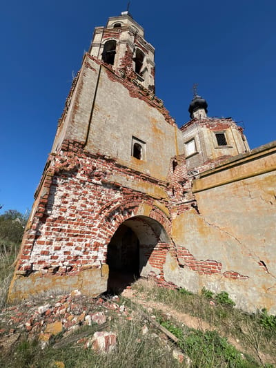 The image shows a dilapidated brick building, partially destroyed and exposed to the elements. The structure appears to be an old church or similar religious building, with a tower visible against a clear blue sky. The location is Шуран, Russia. The building's walls are crumbling, revealing the underlying brickwork, which is a mix of red and white. A large arched opening is visible in one section of the wall, with the surrounding area overgrown with vegetation. The overall impression is one of abandonment and decay.