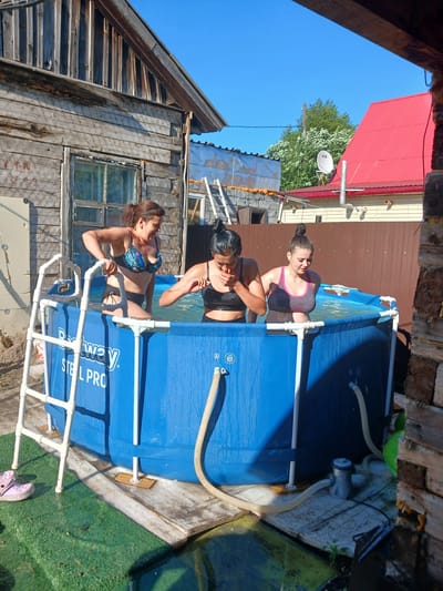 In Chik, Russia, on a sunny day, three young women are in a blue, above-ground swimming pool. The pool is labeled "Bestway Steel Pro." One woman, with brown hair in a ponytail, wears a floral bikini and is standing, reaching for the pool's edge. Another woman, with dark hair pulled into a bun, wears a black sports bra and black bottoms, her face partially obscured as she appears to be splashing water. The third woman, with her hair in a bun, wears a grey and pink sports bra and dark bottoms, looking down into the water. The pool is set up on a concrete base with wooden supports. A white ladder provides access to the pool. In the background, a rustic wooden building stands to the left, with a weathered facade and a window. To the right, a brown wooden fence separates the property from other structures, including a building with a bright red roof and a satellite dish. Green grass and some overgrown vegetation are visible around the pool area. A small water pump with hoses is connected to the pool.