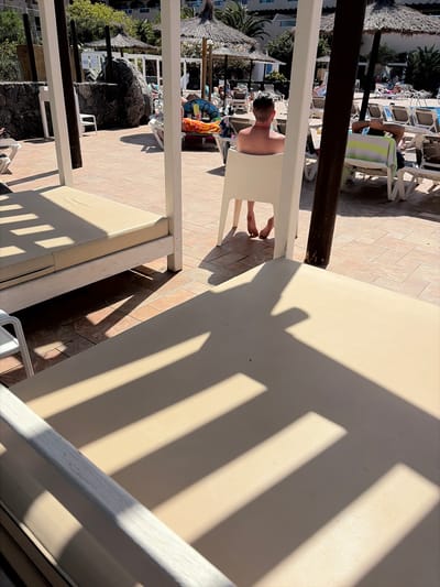 This media file depicts an outdoor scene at a resort, possibly in Antigua, Spain, with strong sunlight casting dramatic shadows from a slatted structure onto a beige surface. The primary subject in focus is a man sitting on a white chair, his back to the camera, with his head and shoulders visible. He appears relaxed, with his arms resting in his lap. A striped towel, green and white, is draped over the chair behind him.

In the background, there are indications of a swimming pool area with loungers and umbrellas. Some people are visible on loungers in the distance, suggesting a daytime leisure activity. The ground is paved with light-colored square tiles. The overall impression is one of a sunny, warm day at a holiday destination.

Notable details include the sharp, linear shadows, indicating direct sunlight, and the presence of what appears to be a thatched umbrella or sunshade on the left side of the image, adding a tropical or Mediterranean feel. The scene is likely set during the daytime, given the strong shadows. No visible text is present in the image. The mood is relaxed and serene, conveying a sense of holiday enjoyment.