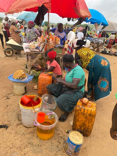 This image captures a vibrant and bustling open-air market scene in Riyom, Nigeria. The setting is a dusty, unpaved area, characteristic of informal markets, with some greenery visible in the distant background under an overcast sky, suggesting it is daytime, possibly late morning or afternoon.

Numerous people, predominantly women, are engaged in commercial activities, acting as both vendors and customers. They are dressed in casual clothing, with many women wearing headwraps and colorful patterned fabrics. The activity centers around buying and selling various goods.

Prominent objects include numerous makeshift market stalls constructed from wood, sheltered by large, colorful umbrellas in red, blue, and white, providing shade from the elements. A wide array of merchandise is displayed, including packaged items, bottles of liquids, spices, and what appear to be cosmetic or personal care products. On the ground, large yellow and orange jerrycans are seen, alongside clear and colored buckets containing liquids, likely cooking oil or palm oil, dispensed with small red plastic cups used as scoops. One bucket holds a pile of golden-brown fried dough balls, a common local snack. A blue and white tin can, possibly containing powdered milk, is also visible. A wheelbarrow is parked near one of the stalls.

Visible text includes "NSDC" on the blue shirt of a man, and "670 N" on a product package, likely indicating a price in Nigerian Naira. The market appears busy and functional, reflecting a typical day of commerce in the region.