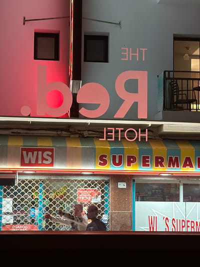 This is a vertical, slightly tilted photograph of a storefront in Sant Antoni De Portmany, Spain. The storefront features a colorful, striped facade with large, pink lettering that reads "THE BG". To the right of "THE BG", the word "HOTEG" is visible in pink letters, followed by "SUPERMA" in red and yellow lettering. The store appears to be a supermarket, as indicated by the text "WI 'S SUPERM" on a white sign to the right of the entrance.

Through a metal grate, two individuals are visible inside the store. One person, wearing a white sweatshirt with a graphic print, is reaching towards a shelf. The other person is partially obscured. Shelves stocked with various products are visible behind them. Red neon lights line the edge of the storefront. Some signs within the store are in Spanish, including "BERIDAS FRIAS" (cold drinks) and "CUANDO QUIERAS" (whenever you want). The overall impression is a brightly lit, possibly bustling, local shop. The time of day is unclear, but the lighting suggests it could be evening or nighttime.