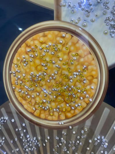 The image shows a close-up view of an ornate decorative piece. The central element is a circular inlay with a golden rim, filled with a textured orange-yellow material that resembles corn kernels. Scattered across this surface are numerous small, clear and yellow rhinestones, adding a sparkling effect. Surrounding this circular inlay, and extending downwards, is a striped pattern in shades of brown and beige. More scattered rhinestones are visible on this striped background, some appearing to be embedded in a glossy finish. The overall impression is of a highly decorative and possibly handmade item, with elements of sparkle and intricate design. The image does not depict any specific location in Giginyu, Nigeria, nor does it contain any people or actions.