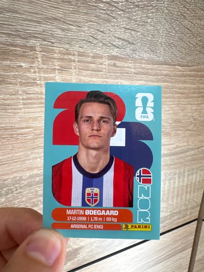 The image is a close-up of a trading card featuring a portrait of Martin Ødegaard, a Norwegian professional footballer. The card has a light blue background with large red and dark blue graphic elements. Ødegaard is depicted wearing a red, white, and blue striped jersey with a crest on the left chest. Text on the card identifies him as "MARTIN ØDEGAARD," with his birthdate "17-12-1998," height "1,78 m," weight "69 kg," and club affiliation "ARSENAL FC (ENG)". The FIFA logo and the year "2023" are visible in the upper right portion of the card. A Norwegian flag symbol is also present. The card is held by a person's hand in the foreground, with their thumb visible. The background consists of a wooden surface with a distinct wood grain pattern. The image is taken from a slightly elevated angle, looking down at the card. There is no visible information or context suggesting a location in Prisovo, Bulgaria.