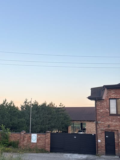 The image captures a section of a brick building with a dark-colored metal gate and a brick wall in front of it. The sky above is clear and transitions from a pale orange near the horizon to a light blue higher up. Three thin power lines stretch horizontally across the upper portion of the frame.

The brick building features a dark brown roof and a window with blinds. To the right of the gate, there is a dark wooden door. The brick wall is topped with decorative brick pillars and has a white sign with a green circle on it. Behind the wall and gate, a line of green trees is visible, with some buildings or structures further in the background.

The scene appears to be a residential or commercial property, likely in a semi-rural or suburban setting. There are no people visible in the image. The lighting suggests it is either early morning or late evening. The location is identified as Obukhivka, Ukraine.