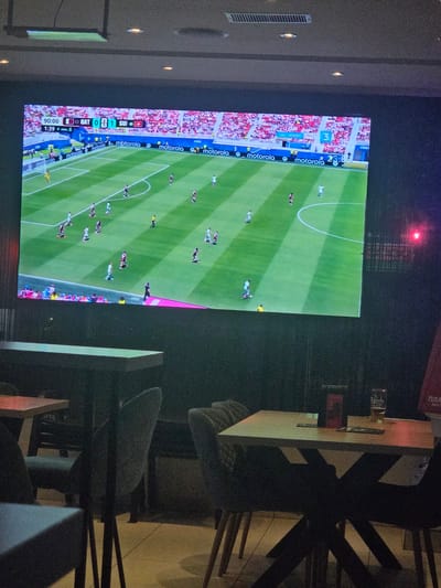 The image shows a large screen displaying a soccer match. The score on the screen indicates "QAT 0 - 1 SUI", with the time at "90:00 + min. 6". Several players in white and red uniforms are visible on the green field, with spectators in the background. The screen is positioned in a dark room, likely a bar or restaurant, as evidenced by the tables and chairs in the foreground. One table has a glass of beer and a menu. The overall lighting suggests an evening setting. The location of Targovishte, Bulgaria, is not discernible from the image itself, and any assertion of it being in Targovishte would require external information.