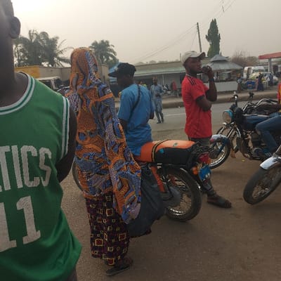 The image captures a daylight scene on what appears to be a paved road or street, likely in an urban or semi-urban setting. The perspective is from a low angle, looking towards a group of people and motorcycles. The sky is bright and slightly hazy.
In the immediate foreground, partially visible on the right, is a person wearing a green sleeveless shirt or jersey with white text. The text clearly shows "CELTICS" and "ICS", suggesting a sports team affiliation, likely "BOSTON CELTICS".
Moving further into the scene, a woman is prominently featured, wearing a long, flowing dress with vibrant and intricate patterns in shades of orange, blue, and brown. She also appears to be wearing a matching head covering. She is standing upright, facing generally towards the left of the frame.
To her right and slightly behind her, a man in a light blue short-sleeved shirt and a dark baseball cap is standing near a motorcycle. Another motorcycle is positioned next to him. Strapped to the rear of this motorcycle is a prominent orange-colored bundle, possibly luggage or goods. A dark, possibly fabric-covered, item is also visible strapped to the side of the motorcycle.
Further to the right, a second man is visible. He is wearing a red short-sleeved shirt and a white baseball cap, standing next to another motorcycle. He is holding an object to his mouth, appearing to be drinking from a bottle or can. This motorcycle also has items strapped to its rear, though less distinctly visible than the orange bundle on the other bike.
In the background, a street extends with other pedestrians sparsely distributed along it. Several low-rise buildings are visible lining the road, and trees, including palm trees on the far left, are scattered throughout the landscape. The overall impression is one of people gathered or waiting by the roadside, possibly for transport or as part of a journey.