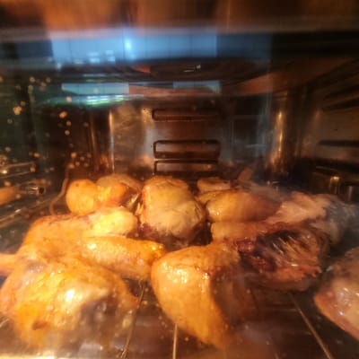 This image shows a close-up view of chicken pieces roasting inside an oven. The chicken is golden brown and appears to be glistening with juices. The oven racks are visible, and there is some condensation on the inside of the oven door, suggesting that the chicken is actively cooking. The background of the image is blurred, but it appears to be the interior of a kitchen. The scene is located in Dunnae-myeon, South Korea, in a domestic setting. There are no people visible in the image.