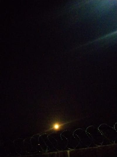 The image is a low-light, slightly grainy photograph taken at night in Mangu Town, Nigeria. The scene is dominated by a dark sky with a faint, hazy illumination from an unseen light source, possibly the moon or a distant streetlamp, positioned in the lower middle portion of the frame. A bright, circular light source, appearing yellow-orange, is visible in the center of the image, casting a soft glow. In the foreground, a coiled barbed wire fence stretches horizontally across the bottom third of the frame. The fence appears to be mounted on a wall, likely made of brick, though the detail is obscured by darkness. The barbed wire itself is faintly illuminated, highlighting its sharp, circular patterns. The overall impression is one of a secured perimeter or boundary, viewed under nocturnal conditions.
