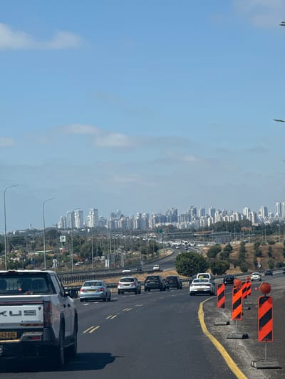 The image shows a highway scene with traffic and city buildings in the background. The highway is paved with asphalt and has yellow lane markings. Orange and black construction barriers are visible on the side of the road. Several cars are driving on the highway, including a white van, a silver car, and a few black cars. A white pickup truck is in the foreground, facing away from the camera. The city in the background is characterized by many tall buildings, suggesting an urban or suburban area. The sky is blue with some clouds, indicating daytime and clear weather. There are power lines and streetlights visible along the highway and in the city. Some vegetation, including trees and dry grass, can be seen on the hills surrounding the highway. The overall impression is a busy highway during the day, likely in a developed area. There is some text visible on the license plate of the pickup truck in the foreground, which appears to be "243-604". The text "LUX" is also visible on the back of the pickup truck.