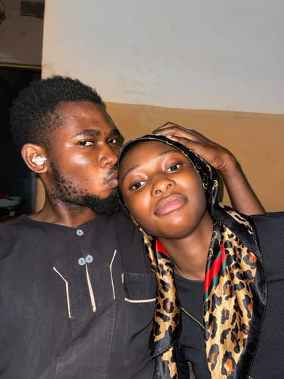 The image features two individuals, a man and a woman, positioned closely together in what appears to be an intimate indoor setting. The image is rotated 90 degrees clockwise, showing the subjects leaning to their right.
The woman is positioned above and slightly behind the man, resting her head on his shoulder or chest. She has dark skin, full lips, and a nose piercing on her right nostril. Her eyes are open, and she is looking directly towards the camera with a calm expression. She is wearing a dark top, and a distinctive black and brown leopard print headscarf or wrap, which also features a red and green striped pattern, is draped around her head and shoulders. Her right hand is visible, resting near her head, possibly on her own headscarf or the man's shoulder.
The man, positioned below the woman, has dark skin, a short beard, and short dark hair. He is looking slightly upward and to his left, seemingly towards the woman or slightly past her, rather than directly at the camera. He is wearing a dark, possibly black or dark blue, collared shirt with visible light-colored stitching details and two light-colored buttons on the front. A white earbud is clearly visible in his right ear.
The background is simple, consisting of plain walls. On the left side of the frame, the wall is a light, possibly off-white or pale gray color. To the right, there's a transition to a darker, beige or light brown colored wall. The lighting appears soft and diffused, typical of indoor lighting.
The overall impression is a warm, affectionate moment captured between the two individuals. There is no visible text in the image.