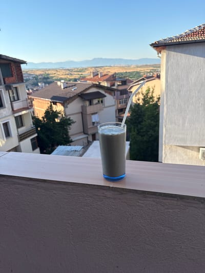 A glass of light-brown liquid with a white straw sits on a light-colored balcony railing, overlooking a city with a backdrop of hazy mountains under a clear blue sky. The city, identified as Sandanski, Bulgaria, features numerous residential buildings with red-tiled roofs. The foreground shows the textured surface of the balcony's lower wall. The image is taken from a slightly elevated perspective.