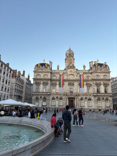 This image depicts the Place des Terreaux in Lyon, France, featuring the Hôtel de Ville de Lyon (City Hall) as the prominent background structure.
The Hôtel de Ville is a large, ornate Baroque-style building with a central clock tower topped by a dome and a spire. Its facade is richly decorated with sculptures, pilasters, and cornices. Two large, vertical rainbow-colored banners, each bearing the text "LYON EST FIÈRE!" (Lyon is Proud!), are suspended from the upper portion of the building, flanking the clock tower. French flags are visible below the banners, above the main entrance. The building's windows are numerous, with several appearing open.
In the foreground, a large, circular stone fountain filled with water occupies the bottom left and center. Several individuals are seated on the fountain's edge, including a person in a red shirt on the right and a person in a yellow top and blue jeans towards the left. To the left of the fountain, an outdoor cafe area is visible with multiple white parasols shading tables and chairs, occupied by patrons.
The square itself is paved with light grey flagstones. Various groups of people are scattered across the square, walking, standing, and interacting. Small ground-level water jets create minor fountains in the mid-ground. Buildings with multiple stories and typical European architectural styles flank the square on the left and right. The sky is clear and light blue, with a few small birds visible in the upper sections. The lighting suggests late afternoon or early evening.