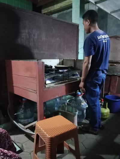 The image shows a man in a blue shirt standing in a room in Kecamatan Brangsong, Indonesia, which is located at the coordinates -6.943 latitude and 110.222 longitude. The man is standing next to a wooden countertop with a gas stove. The countertop is made of wood and has a red stain. There are two empty frying pans on the stove, and a gas tank to the right. The man is looking down at the stove. There are several water jugs and other miscellaneous objects in the room. There is a brown plastic stool in the foreground. The room is dimly lit.