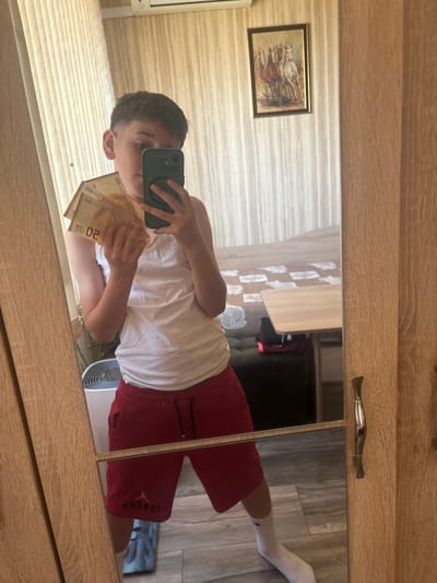 A young person is standing in front of a mirror, holding several euro banknotes in their left hand and a smartphone in their right. They are wearing a white tank top and red shorts, with white socks and sneakers visible in the mirror's reflection. The background of the room includes a wall with vertical striped wallpaper, a framed picture of horses, and a wooden dresser or closet with a decorative handle. The floor appears to be made of grey tiles. The image is a selfie, taken by the person in the mirror. The location is the city of Kazanlak, Bulgaria.