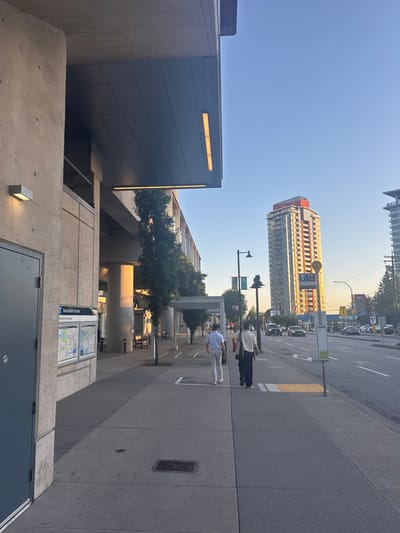 This is a medium-shot, eye-level, outdoor, daytime photograph captured from a sidewalk. The scene depicts an urban environment in Coquitlam, Canada, likely during late afternoon or early evening due to the warm lighting. In the foreground on the left, a concrete structure with a large grey door is visible. Adjacent to the door is a sign for "Burquitlam Station" with a map. The sidewalk extends into the middle ground, where two individuals are walking away from the camera. The person on the left is wearing a light blue shirt and light-colored pants, while the person on the right is wearing a beige shirt and dark pants, carrying a large tote bag. Beyond the sidewalk, a road with moderate traffic is visible. In the background, a tall, multi-story residential building with a red and orange facade dominates the skyline, along with other urban structures and trees. The sky is clear and blue.