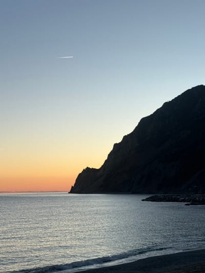 The image displays a coastal scene in Monterosso al Mare, Italy, during twilight, characterized by a clear sky. The upper left portion of the sky exhibits an orange glow near the horizon, transitioning upwards and to the right into a pale blue, with a single white aircraft contrail visible in the upper-middle left. The sea, occupying the lower half of the frame, is calm with subtle ripples, appearing a light blue-grey. Two dark, small buoys are present in the mid-ground water on the left. A large, steep, dark, and heavily shadowed rocky mountain face dominates the right side of the image, extending from the mid-right down to the water's edge. At the base of this mountain on the lower-right, a dark, rocky breakwater or jetty extends into the sea. The immediate foreground shows a narrow strip of dark-colored beach, likely pebbles or coarse sand, with small waves breaking upon it. No people or direct human activity are visible.