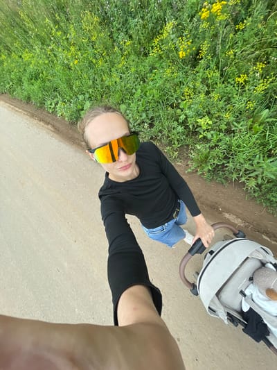 The image shows a person pushing a stroller along a dirt path next to a field of green vegetation with yellow wildflowers. The person is wearing a black long-sleeved shirt, denim shorts, and white sneakers. They are also wearing large, reflective sunglasses with orange and yellow lenses. The stroller is light gray with a brown handle and appears to contain a stuffed animal. The perspective is from the person's point of view, as they are holding the camera in their outstretched arm. The scene appears to be outdoors in a rural or semi-rural setting, possibly on the outskirts of the village of Podshivalovo, Russia, given the context.
