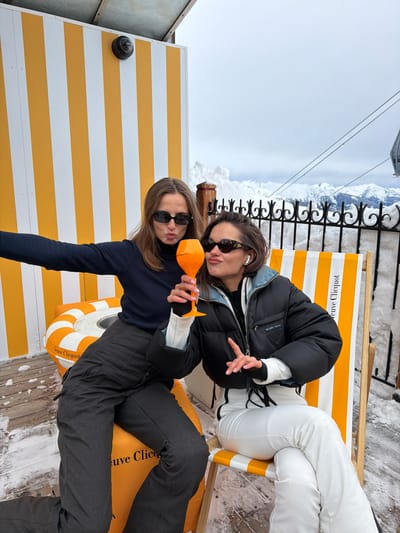 The image captures two young women posing for a photo in an outdoor, snowy, mountain setting, likely a ski resort or a high-altitude lounge area. The overall impression is one of a relaxed, fun social occasion.
In the foreground, to the left, is one woman with long, straight brown hair, wearing dark, rectangular sunglasses, a dark long-sleeved top, and dark pants. She is holding an orange-tinted, uniquely shaped glass, possibly a champagne flute, with a pointed base. Her lips are slightly pursed in a playful pout as she looks towards the camera. She appears to be sitting or leaning on an inflatable, round orange and white striped cushion or float, which has the brand "Veuve Clicquot" printed on it in black lettering.
Behind and slightly to the right of her is the second woman. She has shorter, light brown hair, possibly styled up or pulled back, and is wearing brown-patterned sunglasses and white earbuds. She is dressed in a black puffer jacket over a white long-sleeved top and white pants. Her right hand is raised, making a gesture that resembles a peace sign or a stylized 'V'. She is seated on a wooden deck chair with yellow and white striped fabric, which also prominently displays the "Veuve Clicquot" brand name.
The background shows a stark, snow-covered mountain landscape under a bright, cloudy sky. A black ornate metal fence runs along the edge of what appears to be a viewing platform or balcony, with significant snow accumulation beyond it. Thin wires, possibly from a ski lift or gondola, stretch across the upper part of the frame against the sky. To the far left, a yellow and white striped wall, matching the branding elements, is visible, featuring a small black dome-shaped fixture, possibly a camera or light. The ground is a wooden deck, partially covered in scattered snow. The bright lighting suggests it is daytime. The women appear to be enjoying themselves and are dressed for cold weather.