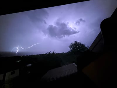 The image presents a nighttime scene in Zornitsa, Bulgaria, depicting a powerful lightning strike illuminating a stormy sky. A brilliant, jagged white lightning bolt extends from the upper left portion of the frame, curving downward towards the horizon. The flash casts a strong purplish-blue hue across the dense, dark cloud formations. Additional illumination is visible within the clouds towards the upper right, suggesting further electrical activity. The viewpoint is elevated, looking out from what appears to be a window or balcony, framed by dark architectural elements along the top and right edges. In the lower-center, the dark silhouette of a corrugated or tiled roof is visible, with a portion of another structure featuring a light-colored section or window to its left. The midground and background reveal a landscape primarily covered by silhouetted trees and foliage. Numerous small, bright lights, consistent with streetlights or distant buildings, dot the expansive, dark landscape extending into the distance. No individuals or discernible actions are present within the frame.