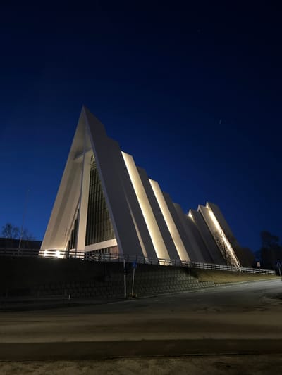The image, captured at night, presents a striking modern building with angular architecture against a deep blue, clear sky. The photo is oriented vertically, showing the building on the left half of the frame, extending from the bottom-left to the upper-right when viewed with the building upright.

The structure is predominantly white, characterized by a series of receding, stepped layers. Integrated warm yellow strip lights illuminate the underside of these layers, casting a bright glow upwards and highlighting the building's geometric form. Its leftmost point forms a sharp, triangular apex.

To the right of the building, a paved pathway or sidewalk, constructed from interlocking bricks, runs alongside. Beyond this, a darker, unpaved or rough road surface is visible. Silhouetted bare tree branches can be seen in the upper right background, contrasting with the illuminated building.

The deep blue-black sky suggests a clear night with no visible clouds. A few extremely faint points of light, possibly stars or distant airborne objects, are discernible in the expansive sky. A standard European-style street sign, featuring a blue square with a white symbol (likely for pedestrians or traffic direction), is mounted on a pole near the paved area on the right.

No people or specific activities are depicted, giving the scene a serene and architectural focus. The setting is an urban or semi-urban environment in Tromsø, Norway.