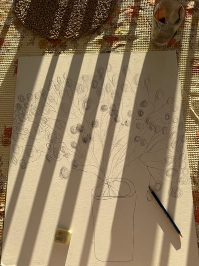 An overhead photograph taken in Kfar Vradim, Israel, captures an artistic workspace under strong, direct sunlight casting distinct, parallel vertical shadows across the scene. The central subject is a textured, off-white canvas featuring a graphite pencil sketch of a stylized plant arrangement in a cylindrical vase. The plant consists of multiple slender stems branching upwards, each terminating in a spiraled, circular form resembling a berry or bud. To the lower right of the canvas lies a blue pencil, while a white eraser with "JOHN SHEN" printed on it is positioned on the lower left. The canvas rests on a light-colored, patterned tablecloth with a floral design and a grid texture. In the upper left corner, a circular woven mat made of natural fibers is partially visible. A ribbed glass tumbler containing dried plant material is situated in the upper right quadrant of the image. No individuals are present.
