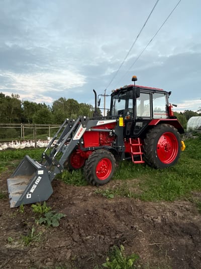 In the rural outskirts of Kukui, Russia, a vibrant red and black tractor with a front-end loader is parked on a patch of uneven, dark brown earth and green grass. The tractor features a fully enclosed cabin with tinted windows, an orange rotating beacon on its roof, and multiple exterior lights. The front loader is lowered, with its grey bucket resting on the ground, partially filled with soil and a small amount of green vegetation. The tractor's large rear wheels are bright red, contrasting with the black tires. In the background, a wooden fence encloses a flock of white turkeys resting on the grass. Beyond the fence, a dense line of green trees forms a natural backdrop. The sky above is overcast with soft, grey clouds, suggesting a late afternoon or evening setting. A single power line stretches across the sky above the tractor.