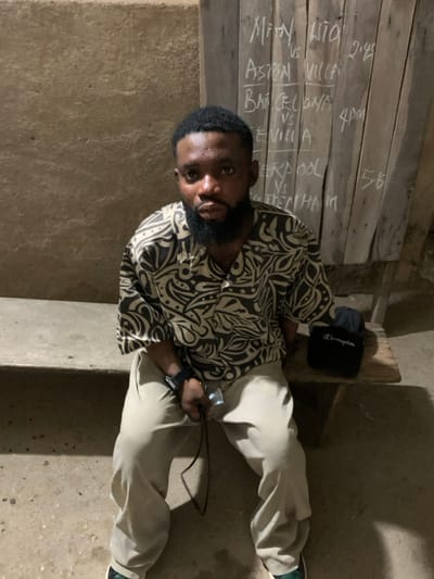 A dark-skinned male individual is seated on a rustic wooden bench in Okene, Nigeria. He wears a short-sleeved collared shirt with a black and light-patterned design, light-colored trousers, and a dark smartwatch on his left wrist. In his left hand, he holds a black cable with a silver metallic end. His gaze is directed forward. To his right, on the bench, rests a black pouch with "Champion" inscribed in white, next to a folded dark fabric item. The background features a textured, light brownish-grey concrete or mud wall on the left. On the right, a vertically planked wooden board displays handwritten chalk text listing football matches and times: "MAN UTD vs ASTON VILLA 2:45", "BARCELONA vs SEVILLA 4pm", and "LIVERPOOL vs TOTTENHAM 5:6". The scene is dimly lit, suggesting an indoor or covered setting.
