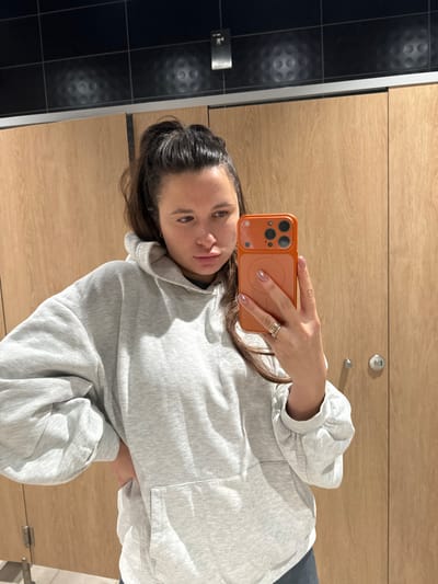 A young woman poses for a selfie in a public restroom. She holds an orange smartphone with her right hand, which has multiple rings on the ring finger. She wears a light grey hoodie with balloon sleeves and a pocket. Dark hair is partially pulled back. The background shows a wall of light woodgrain paneling and a section of dark, textured tile.