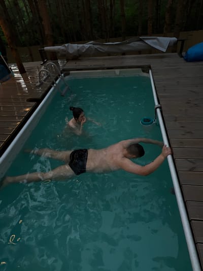 In Ivachiv Horishnii, Ukraine, two individuals are present in an outdoor swimming pool. The pool is rectangular and surrounded by a wooden deck. A male, wearing dark swim trunks, is floating on his back in the foreground, with his arms extended. A female, wearing a light-colored bathing suit, is partially submerged in the water further back in the pool, with her hands near her face. The water is a clear turquoise color and appears to be rippling. The environment is dusky, with trees visible in the background. A pool cover is draped over a structure to the left of the pool.