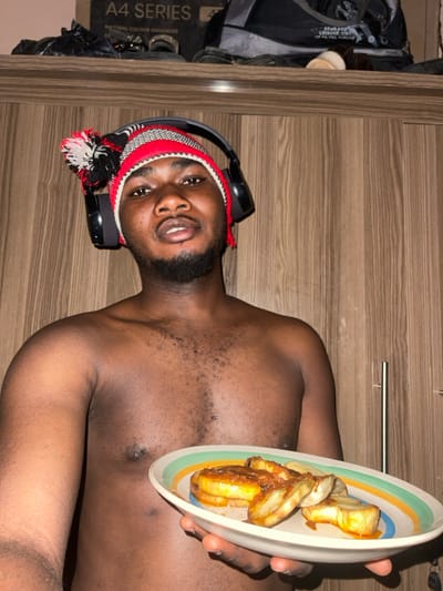 An adult male stands shirtless in what appears to be an indoor setting in Unguwar Gabas, Nigeria. He is positioned centrally, looking directly at the camera with a neutral expression and slightly parted lips. The man has a dark complexion, short dark hair, and a visible beard and mustache. He wears a red, black, and white striped knitted beanie with a prominent pom-pom, over which black over-ear headphones are placed. His left hand holds an oval-shaped white plate with green, orange, and light blue stripes on its rim. The plate contains several slices of fried plantain or yam, topped with a reddish-brown sauce. The background features light brown wooden paneling, likely a wardrobe or wall, with visible vertical wood grain. Above the man's head, on a shelf, a black box labeled "A4 SERIES" and various items including shoes and a grey bag are visible. A metallic handle is partially visible on the right side of the wooden paneling.