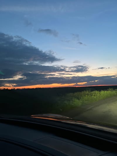 The image depicts a rural landscape at dusk, likely captured from a vehicle. The sky is a gradient of blues and oranges, with scattered clouds. The horizon is lined with a dark silhouette of trees against the vibrant sunset. In the foreground, the interior of a car is visible, including the dashboard and a windshield reflecting the twilight sky and surrounding greenery. The fields are dark, with some areas illuminated by what appears to be headlights or artificial lighting, casting a green glow on the vegetation. The location is identified as Poltavka, Ukraine, suggesting an agricultural or undeveloped area. There are no people or animals visible in the scene. The overall mood is tranquil and serene, characteristic of a sunset in a rural setting.