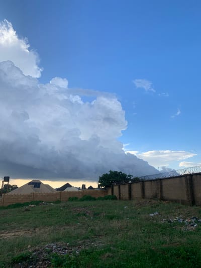 The image depicts an outdoor scene in Rafin Bauna, Nigeria, with a focus on the sky and the immediate surroundings. The sky is a dominant feature, with a bright blue expanse on the left transitioning into a dramatic display of large, white, and grey clouds that occupy the center and right of the frame. The clouds suggest impending weather, possibly rain or a storm. The lighting indicates it is either early morning or late afternoon, with the sun casting a warm glow behind the clouds on the horizon.
In the foreground and along the right edge of the image, a grassy area is visible, interspersed with some scattered debris and a concrete wall topped with barbed wire, suggesting a boundary or enclosure. Behind the wall, the rooftops of several buildings are visible, some with what appear to be solar panels. A single tree with green foliage stands near the wall. The scene is still and quiet, with no people visible. The overall impression is one of a natural landscape under a dynamic sky, with human elements present but not the primary focus.