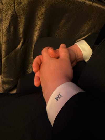 A close-up shot showcases two hands, clasped together. The person on the right wears a dark suit jacket with a crisp white cuff visible, adorned with the initials "IM" in a dark font. The other hand is also fair-skinned. The texture of a dark, possibly velvet, material is seen in the bottom left corner of the image. The lighting is subdued.