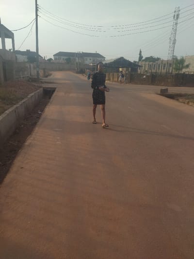 The image shows a woman standing on a paved road in New Karu, Nigeria, at the coordinates 8.981 latitude and 7.559 longitude. She is wearing a black dress with sequins and is looking directly at the camera. The road is empty and there are no cars or other people in the image. On the left side of the image, there is a concrete curb and a grassy area. On the right side of the image, there are a few buildings in the distance. The sky is overcast and there are power lines running across the image. The image appears to be taken in the early morning hours. The scene is characterized by emptiness, quiet, and tranquility. The single person present is the woman who is the focus of the image. The woman stands in the middle of the empty road, her stance and posture suggesting a sense of calm composure. The surrounding environment is characterized by low-rise buildings, concrete structures, and a dusty, brown road. The light is soft and diffused, giving the image a hazy and ethereal quality. The overall impression is one of quiet solitude and a sense of waiting.