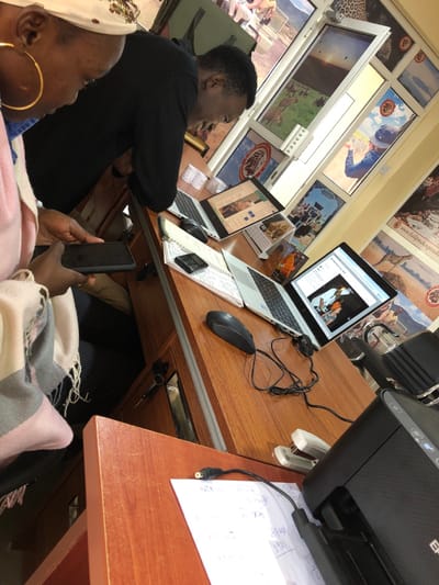 The image shows a scene inside an office in Arusha, Tanzania, located at coordinates -3.372 latitude and 36.689 longitude. Two people are seated at a wooden desk, facing each other. The person on the left is an adult female wearing a pink and grey patterned scarf and a head wrap. She is holding a black smartphone. The person on the right is an adult male wearing a black shirt. He is looking at a laptop. There are three laptops on the desk, one of which is open. The open laptop has a screen displaying a webpage with a photo of two people in a safari vehicle. Several open notebooks and a black calculator are on the desk. A black computer mouse is on the desk in front of the male. There is a black printer on the desk in the lower right corner. The office walls are cream-colored with windows. Several framed posters are hanging on the walls, showing pictures related to safari tourism. The posters on the right are partially obscured by the printer. The scene is brightly lit.