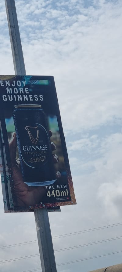 An outdoor Guinness advertisement is displayed on a pole against a cloudy sky. The advertisement prominently features a can of Guinness Foreign Extra Stout held in a hand. The text "ENJOY MORE GUINNESS" is visible at the top of the ad, with "THE NEW 440ml" and the website "DRINKIQ.COM" at the bottom. The advertisement is located on a pole in Ikoyi, Nigeria, with power lines visible in the background.