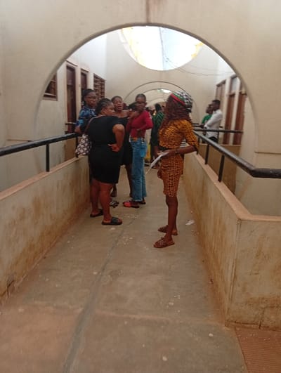 The image shows an arched concrete walkway in Nsukka, Nigeria. The passageway features a light-colored concrete floor with minor stains, and low concrete barrier walls topped with black metal railings extend along both sides. Windows with security grilles and wooden doors are visible along the left wall. The ceiling consists of multiple arches, and natural light enters from above and through openings.
Approximately nine individuals are present within the walkway. On the left, a group of five women stands together. One woman wears a black dress and black sandals, standing with her left hand on her hip. Another woman behind her wears a blue patterned top and eyeglasses. A third woman wears a red long-sleeved top, blue jeans, and red slides.
In the right foreground, a woman in an orange and black patterned short-sleeved dress, brown slides, and a red, green, and black patterned head scarf stands facing left, holding white papers in her left hand. Further down the walkway on the right, two men are visible near the railing; one wears a white long-sleeved shirt, and the other a green shirt. The individuals appear to be standing and conversing in small groups within the passageway.