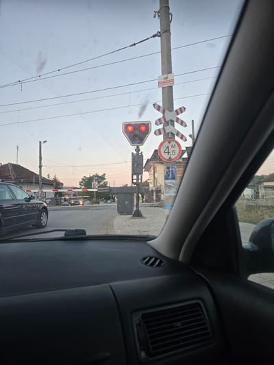 The image depicts a view from inside a vehicle at a railroad crossing in Slivnitsa, Bulgaria. The crossing gate is down, indicated by the red signal lights and the striped barrier arms. A railroad crossing sign with red and white diagonal stripes is prominently displayed on a pole. Below it, a circular sign shows "4.5 M," likely indicating a height restriction. Another sign on the pole, partially obscured by glare, appears to be a warning sign with text and a camera symbol, suggesting surveillance. Smoke can be seen rising from a distant source, possibly from a train or industrial activity. The sky is overcast with a pale hue, and overhead electrical wires are visible, characteristic of an electrified railway line. In the background, buildings with pitched roofs line the street, and other vehicles are stopped at the crossing. The interior of the vehicle shows a portion of the dashboard and windshield.