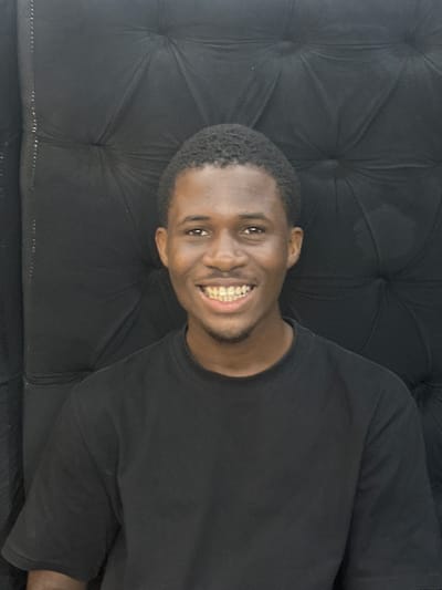 A young African male with short hair and a black shirt is smiling at the camera. He appears to be sitting on a black, tufted couch or upholstered surface. The location is unknown.