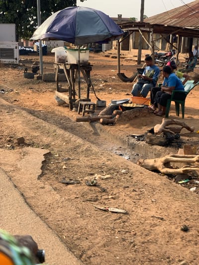 The image depicts an open-air, roadside scene in Kabba, Nigeria, characterized by an unpaved, reddish-brown dirt ground with a shallow drainage furrow running horizontally across the midground.
In the mid-ground, a makeshift food stall is present, covered by a blue and grey striped umbrella providing shade. The stall consists of a wooden frame holding two transparent plastic containers, one with a green lid, likely containing food items. A small wooden stool is positioned near the base of the stall.
Adjacent to the stall, to the right, three individuals are seated around an open-air cooking setup. An active fire, fueled by logs, burns beneath a metal pot or pan containing yellowish food items. A male, wearing a blue patterned t-shirt and jeans, sits on a light-colored plastic chair, looking down at and manipulating a mobile phone. To his right, a female, dressed in a blue t-shirt and dark trousers, is seated on a green plastic chair, observing the cooking area. A third person, partially obscured, is seated further to the right on another green plastic chair.
The ground around the cooking area and the drainage furrow contains scattered debris, including charred wood, ash, plastic litter, and a knife with a light-colored handle. In the background, multiple single-story buildings with corrugated metal roofs are visible, along with a parked motorcycle and a repurposed tire. To the far left, an external white unit and stacked pipes are visible. The scene is illuminated by natural daylight, casting subtle shadows.
